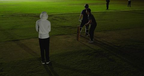 Night cricket with spectator in white hoodie and bucket hat watching batsman at stumps