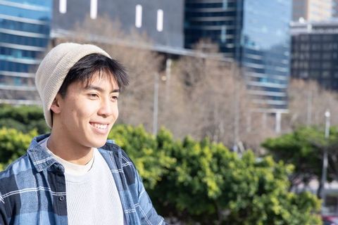 Sunlit urban teen smiling while standing in city greenspace wearing beanie and plaid shirt