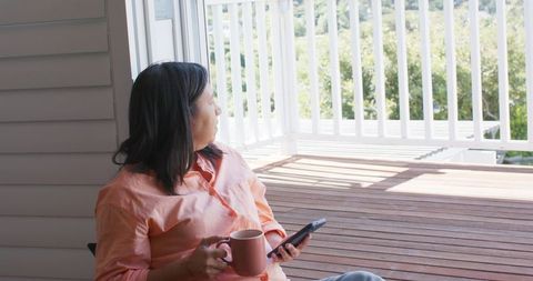Mature asian woman sitting on sunlit porch holding mug and phone gazing at garden view