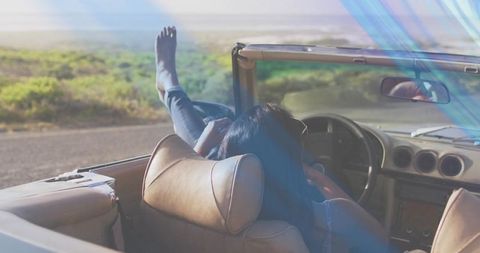 Indian woman relaxing in convertible with feet up on coastal road during golden hour