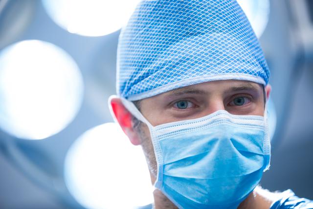 Close-up of a surgeon in an operation room, wearing a surgical mask and cap. Ideal for use in medical and healthcare-related content, articles about surgery, hospital environments, and medical professionals at work.