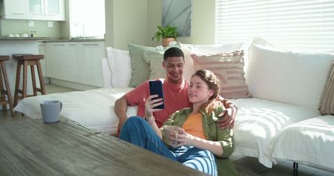 Couple relaxing on floor sharing smartphone and coffee in sunlit modern living room