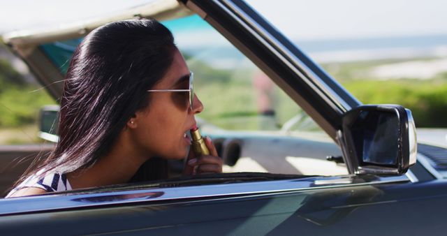 Depicting a young woman putting on lipstick while seated in a convertible car, with a beach in the background. The setting is sunny and outdoors, epitomizing a carefree summer day. The woman wears sunglasses, indicating a stylish look. Perfect for advertisements related to cosmetics, summer vacations, travel, car commercials, or lifestyle blogs focusing on beauty and leisure.