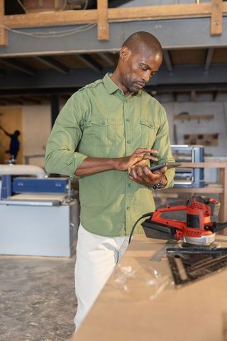 Mature african american craftsman using tablet at woodworking bench with power sander