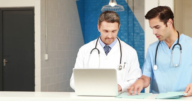 Two healthcare professionals reviewing medical records using a laptop in a modern hospital. One doctor in a white coat is intently focused on the laptop, while the other in blue scrubs is pointing at written notes. Ideal for illustrating teamwork in healthcare, digital record-keeping, modern medical practice, and professional collaboration in medical settings.