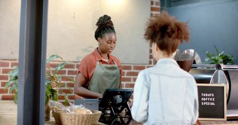 African american barista serving customer in rustic brick coffee shop operating pos terminal