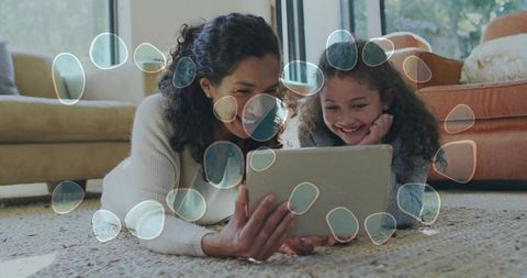 Mother and daughter lying on carpet watching tablet, sharing smiling family bonding moment