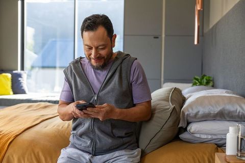 Asian man using smartphone while sitting on bed edge in modern cozy bedroom with natural light