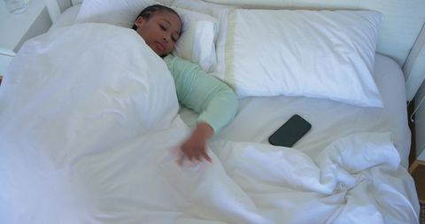 African american woman reaching for smartphone while resting in bed under white duvet