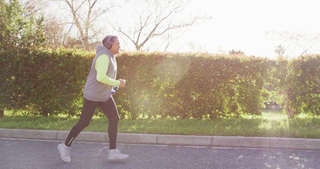 Senior man dressed in sport outfit jogging along a suburban road in the park during sunlight. Ideal for illustrating active lifestyles, senior health and fitness, morning exercise routines, and outdoor activities for elders.