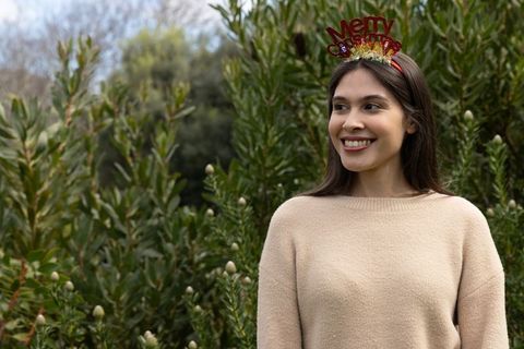 Young woman wearing merry christmas headband smiling amid lush green foliage, festive