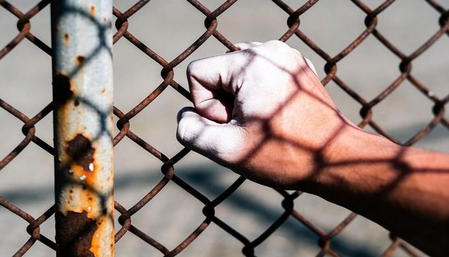 Clenched fist pressing rusted chain-link fence casting geometric shadow on forearm