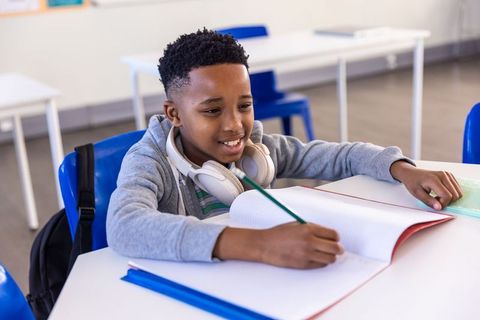African american boy writing in notebook at school desk wearing headphones, concentrating