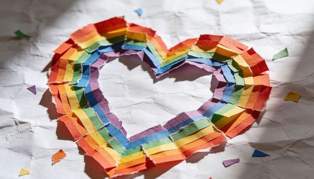 Showing layered rainbow heart torn paper strips on crumpled white sheet with confetti