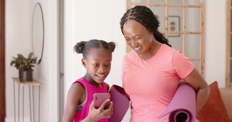 African american mother and daughter preparing for home yoga, smiling and checking phone