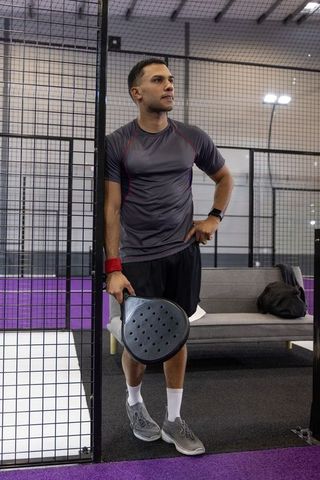 Leaning against metal fence holding paddle on indoor padel court after match