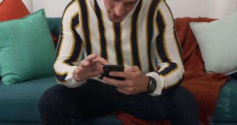 Young man leaning forward on sofa using smartphone, striped sweater and smartwatch