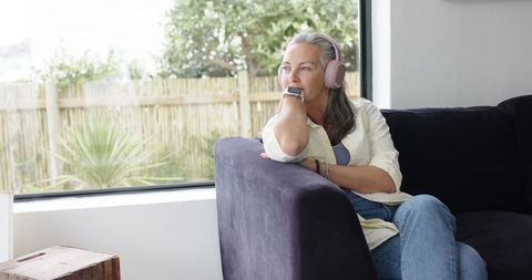 Mature woman relaxing on couch by window wearing pink headphones and smartwatch, contemplative
