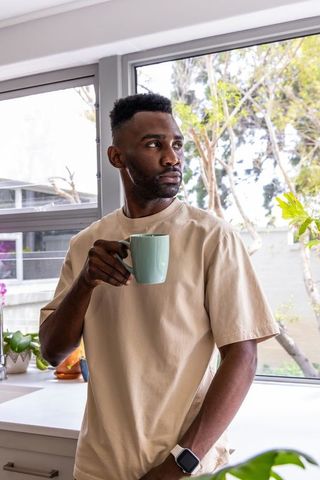 Contemplative african american man standing in sunlit kitchen holding mug, gazing outside