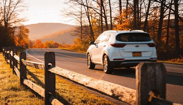 White suv driving along scenic autumn road by wooden fence at golden hour sunset