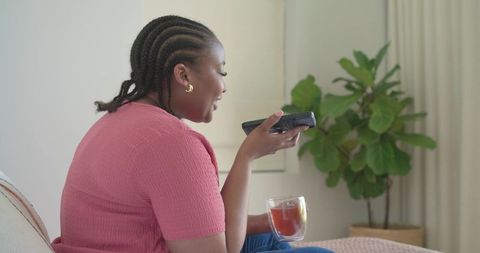African american woman speaking into remote while sipping tea on sofa with houseplant