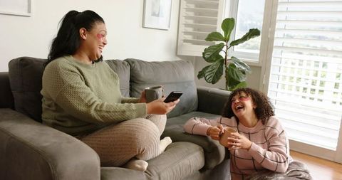 Friends laughing on grey sofa holding mugs and smartphone during cozy morning at home
