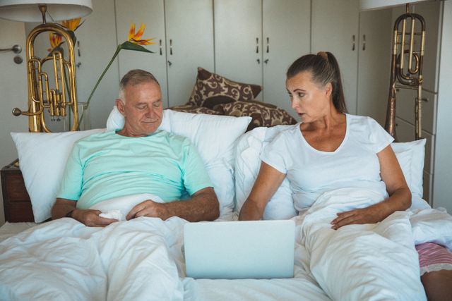 Senior couple lying in bed, using a laptop together. They are wearing pyjamas and appear relaxed and comfortable. This image can be used for topics related to elderly lifestyle, technology use among seniors, home quarantine, and family bonding.