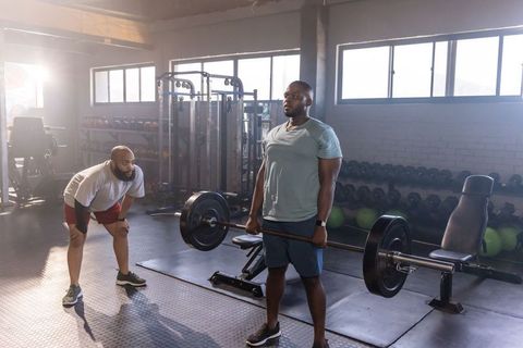 African american man deadlifting while trainer spotting in light-filled strength gym