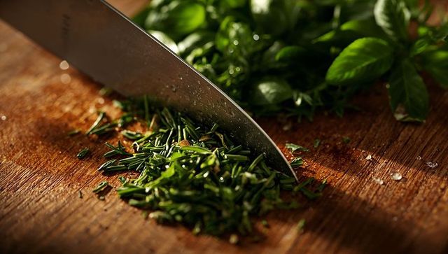 Chopping fresh rosemary and basil with stainless-steel chef knife on wooden cutting board
