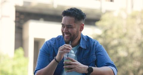 African american man smiling while enjoying green smoothie outdoors in denim jacket