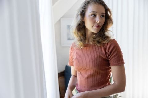 Contemplative young woman standing by bright window wearing ribbed rust top in minimal home