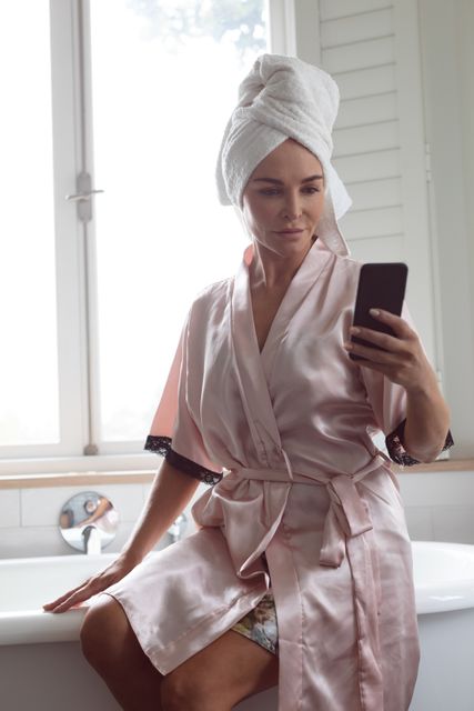 Woman sitting on edge of bathtub in bathroom, wearing bathrobe and towel on head, taking selfie with smartphone. Perfect for themes related to self-care, morning routines, technology use, and casual home lifestyle.