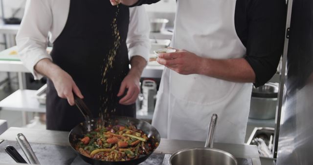 Chefs preparing a stir-fry dish in a professional kitchen setting, demonstrating teamwork and culinary skills. One chef is stirring the vegetables in a frying pan while the other is adding herbs or spices. This imagery is perfect for articles on culinary arts, restaurant advertising, cooking classes, and professional chef profiles.