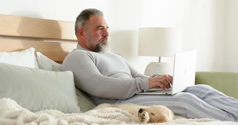 Mature man working on laptop in bed with sleeping puppy in cozy minimalist bedroom