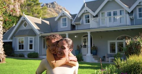 Mother carrying daughter piggyback smiling on lush lawn in front of gray two-story home