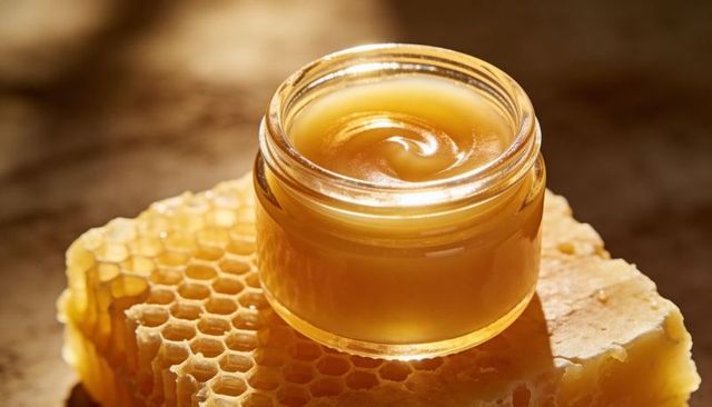 Swirling creamy honey in small glass jar on natural beeswax honeycomb macro texture