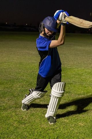 Teen cricketer batting under floodlights at night wearing helmet and pads on grassy pitch