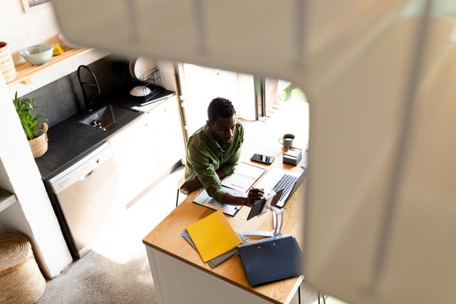 High angle view of an African American man working from home in a kitchen. He is using a laptop and a tablet, surrounded by paperwork and folders. This image is ideal for illustrating concepts related to remote work, home office setups, modern lifestyles, and productivity. It can be used in articles, blogs, and advertisements focusing on work-from-home trends, technology in daily life, and inclusive work environments.