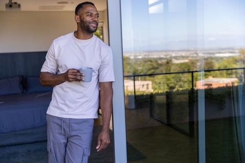Mature african american man leaning on sliding door holding mug, balcony city view