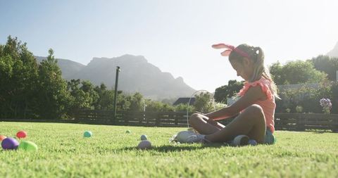 Young girl wearing bunny ears sitting on lawn collecting colorful easter eggs in backyard