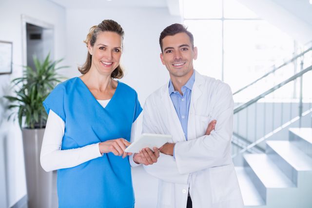 Nurse and doctor standing in hospital corridor using digital tablet, smiling at camera. Ideal for healthcare, medical technology, teamwork, and patient care concepts. Useful for medical websites, hospital brochures, and health-related articles.