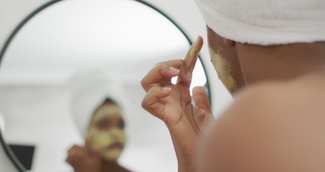 This image depicts a woman applying a facial mask in front of a mirror with a towel wrapped around her head. She appears focused on her beauty routine, highlighting the importance of self-care and skincare. This image is ideal for use in articles or advertisements related to beauty, wellness, and skincare products. It can also be utilized in social media posts promoting self-care practices and natural skincare routines.