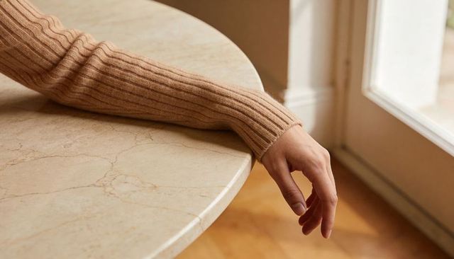 Relaxed forearm and hand resting on beige marble table with ribbed sweater sleeve
