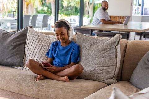 Father working remotely while teen son listening to music on headphones using smartphone