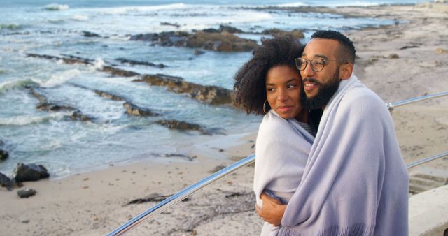Romantic couple wrapped in blanket, embracing as they enjoy tranquil seaside view, highlighting themes of love and connection. Ideal for promoting travel destinations, relationship advice, lifestyle blogs, and cozy outdoor clothing.