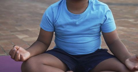 Child meditating cross-legged on purple mat on brick patio wearing blue shirt and shorts