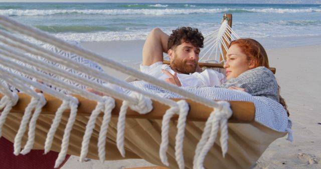 A young Caucasian couple relaxes in a hammock on the beach, enjoying a serene moment together, with copy space. Their closeness and peaceful surroundings suggest a romantic getaway or a tranquil vacation moment.
