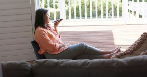 Mature woman relaxing on floor by open doorway, speaking into smartphone on sunlit deck