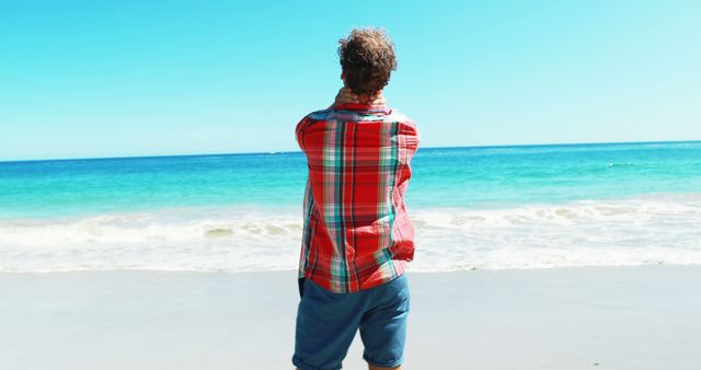 Man standing on sandy shoreline, facing ocean. He wears a casually chic outfit featuring a bold plaid shirt and shorts. Bright blue sea and clear sky evoke tranquility, ideal for travel blogs, vacation ads, lifestyle content, or social media posts promoting beach destinations.