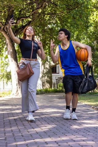 African american friends taking selfie while walking with basketball in leafy suburb park